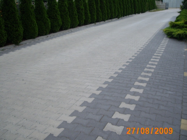 Szeroki podjazd wyłożony jasnoszarą kostką brukową o nieregularnym kształcie, z dekoracyjnym pasem ciemnoszarej kostki z jasnymi wstawkami po prawej stronie, obrzeża z zielonych krzewów i wysokich...